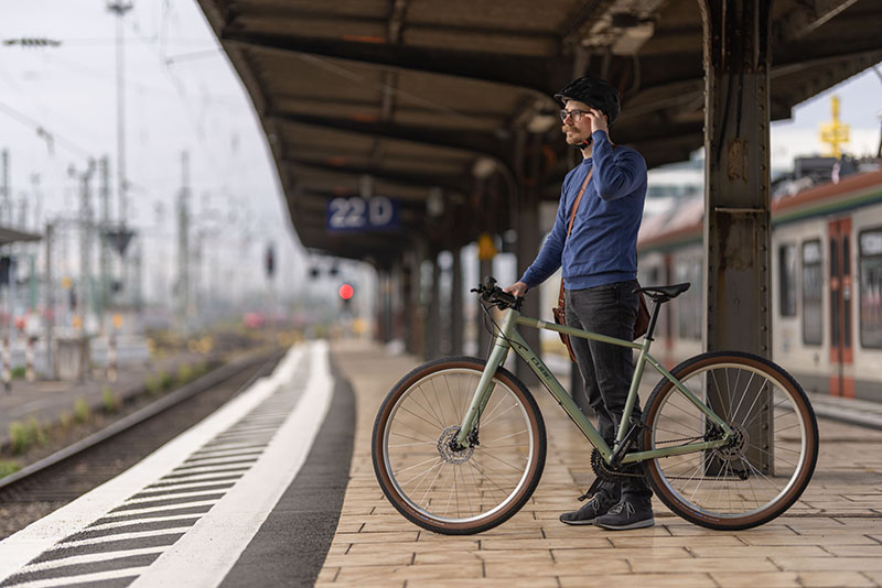 Ein Hipster-Tpy-Mann steht mit seinem olivgrünen Stadtrad an einem Bahngleis und hält sich am Kopf. Er trägt einen Schnauzbart und einen Helm.