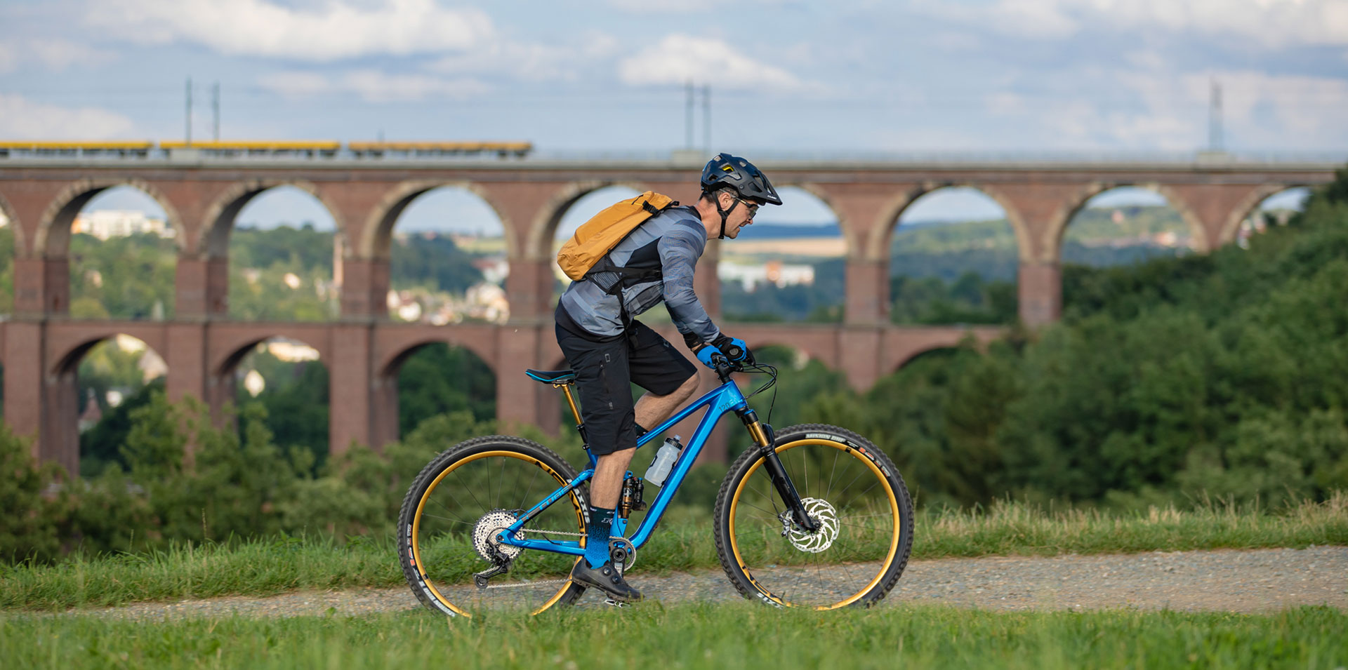 Ein Mountainbiker mit gelbem Rucksack fährt vor Landschaft stehend auf seinem Rad. Im Hintergrund ist groß ein Viadukt zu sehen.