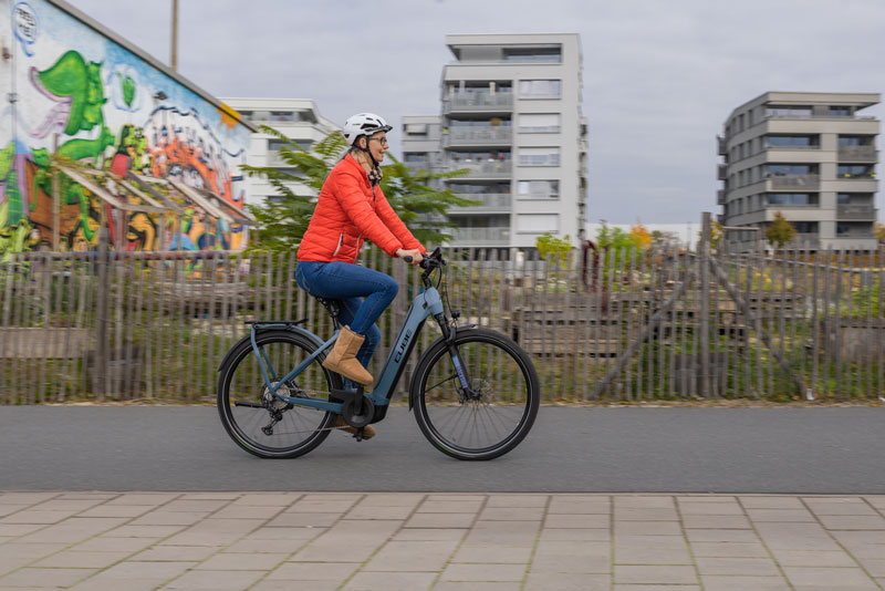 Eine Radfahrerin im Rentner-Alter fährt auf einem grauen E-Bike der Marke Cube durch ein Stadtgebiet. im Hintergrund sind Hochhäuser, Gärten und eine mit bunten Graffiti besprühte Wand.